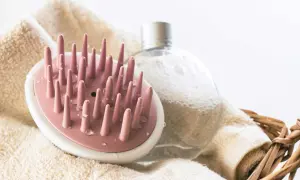 Rosa borste för att exfoliera hårbotten, bredvid en bubblig flaska och en handduk i en korg.