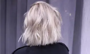 En person med axellångt, texturerat ljusblont hår står framför en grå fond.