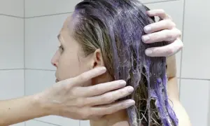 En person masserar in silverschampo med blå pigment i sitt blonda hår. Håret blir lila där schampot appliceras.