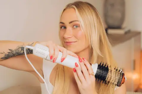 Blond kvinna med tatuering på armen använder en stylingborste för att skapa volym och glans i sitt blow out hår.