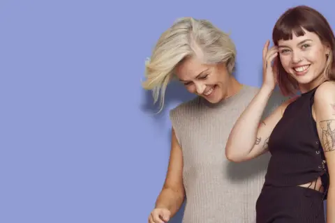 Två kvinnor ler mot kameran. Den ena har en kort rufsig frisyr och grått hår, den andra har mörkt hår och tatueringar.