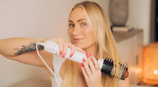 Blond kvinna med tatuering på armen använder en stylingborste för att skapa volym och glans i sitt blow out hår.