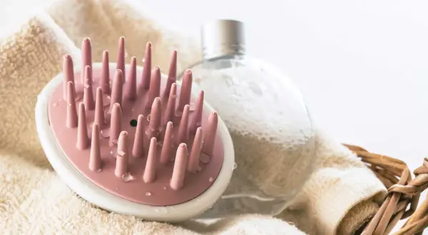 Rosa borste för att exfoliera hårbotten, bredvid en bubblig flaska och en handduk i en korg.