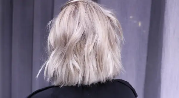En person med axellångt, texturerat ljusblont hår står framför en grå fond.