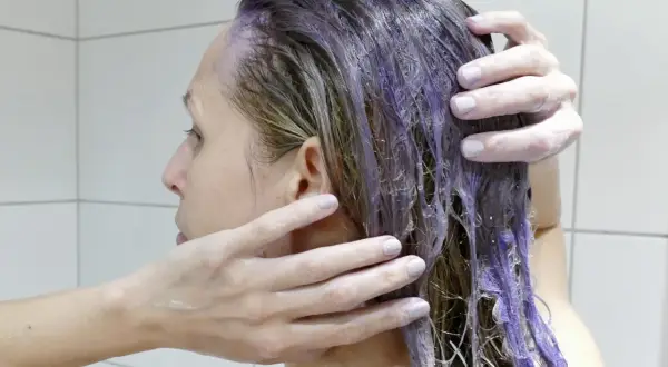 En person masserar in silverschampo med blå pigment i sitt blonda hår. Håret blir lila där schampot appliceras.