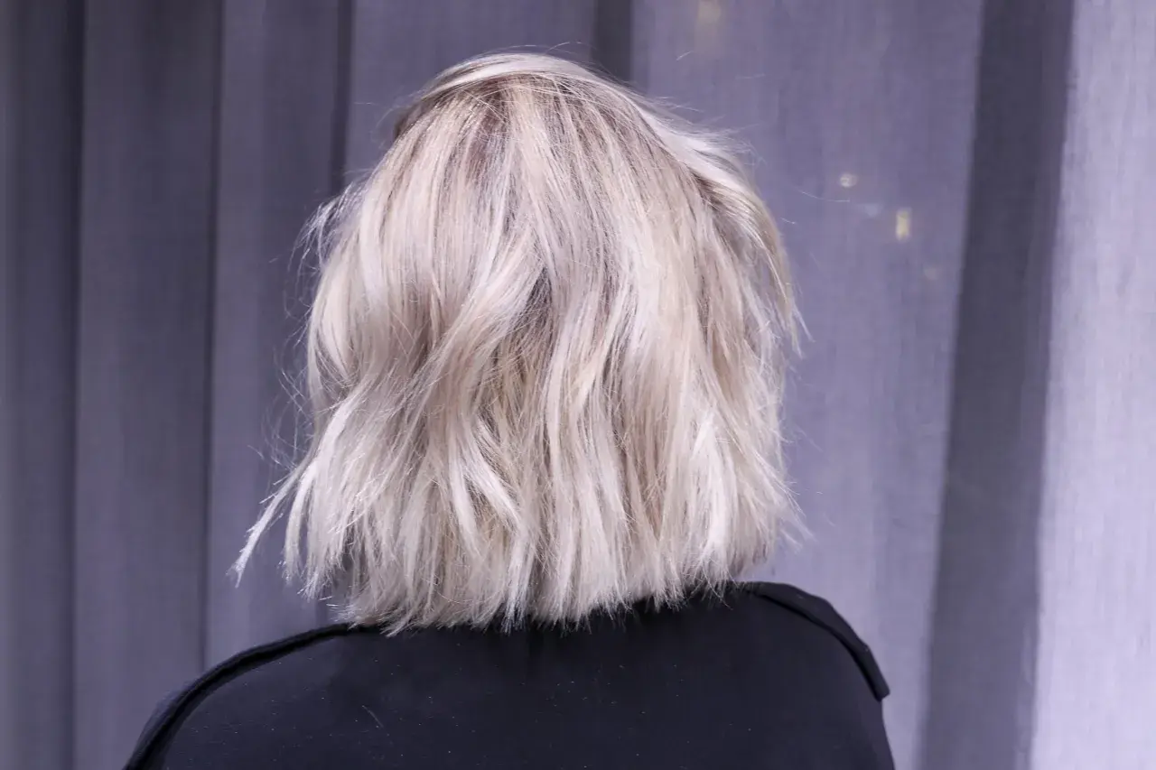 En person med axellångt, texturerat ljusblont hår står framför en grå fond.