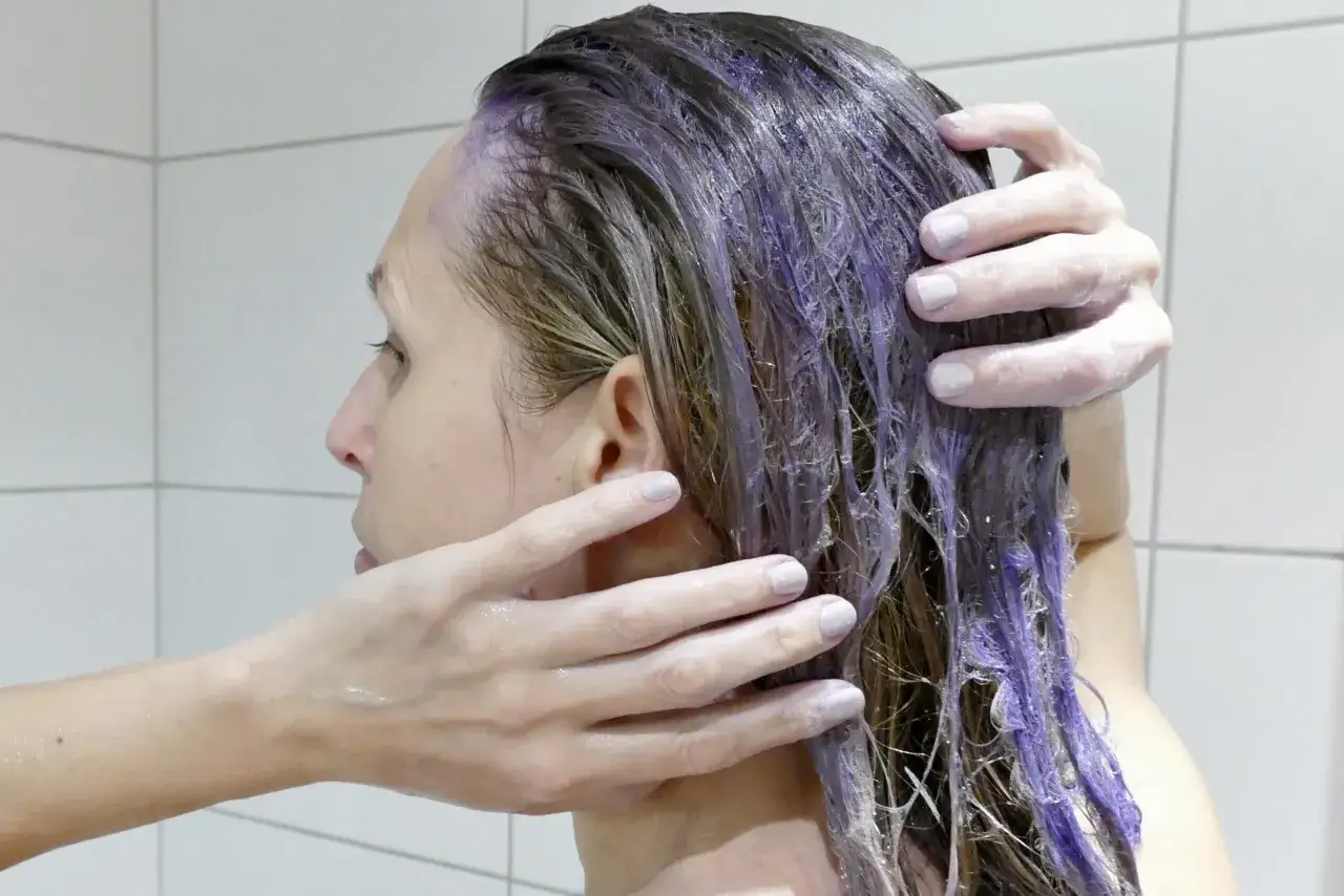 En person masserar in silverschampo med bl&aring; pigment i sitt blonda h&aring;r. H&aring;ret blir lila d&auml;r schampot appliceras.