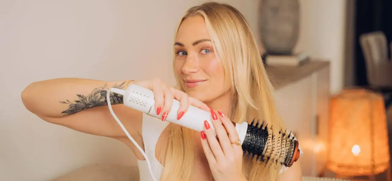 Blond kvinna med tatuering p&aring; armen stylar sitt h&aring;r med en varmluftsborste f&ouml;r perfekt blow out h&aring;r.