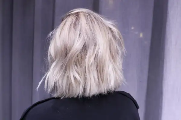 En person med axellångt, texturerat ljusblont hår står framför en grå fond.