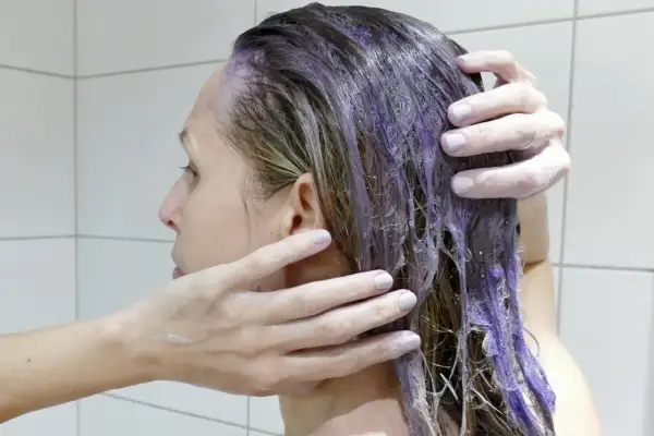 En person masserar in silverschampo med blå pigment i sitt blonda hår. Håret blir lila där schampot appliceras.